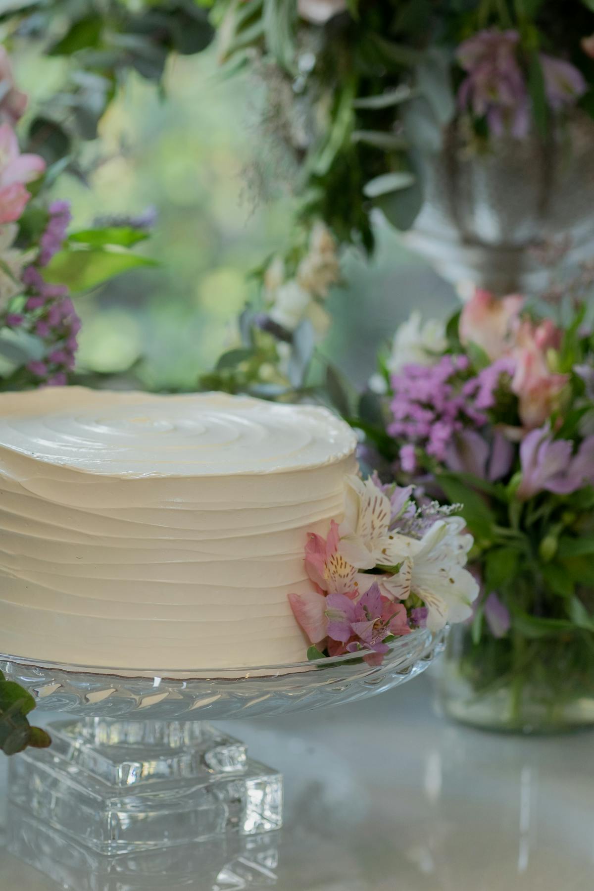 A white buttercream celebration cake on a glass stand, dressed with fresh alstroemeria and pink florals