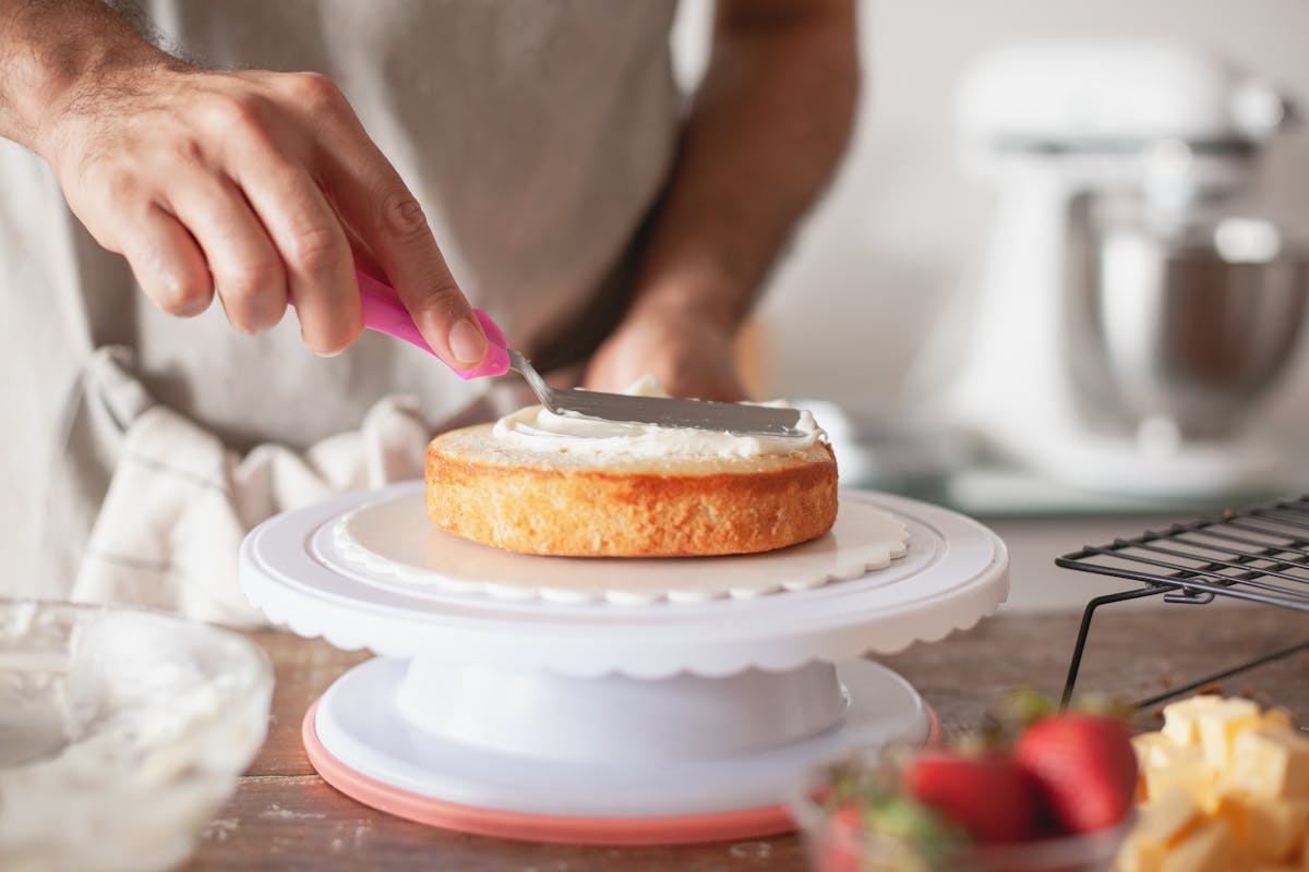 A baker's hands smoothing buttercream icing onto a sponge cake with a palette knife