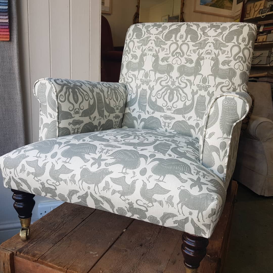 A traditional armchair reupholstered in a striking blue-grey patterned fabric featuring birds, beasts and folk-art animals — antique frame on dark turned legs and brass castors